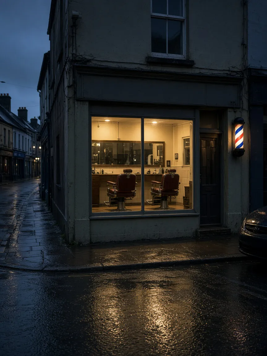 The shop on a corner at dusk: a lit window with two empty red leather chairs, a striped pole on the wall, and the wet street in front.