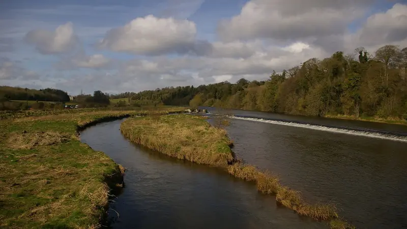 The River Boyne curving past a low weir, with trees on the far bank under a broken blue and white sky
