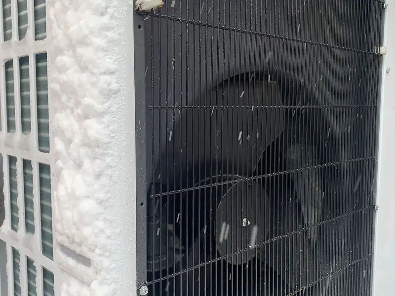 The outdoor fan unit of an air-to-water heat pump on a brick wall, frosted over with snow