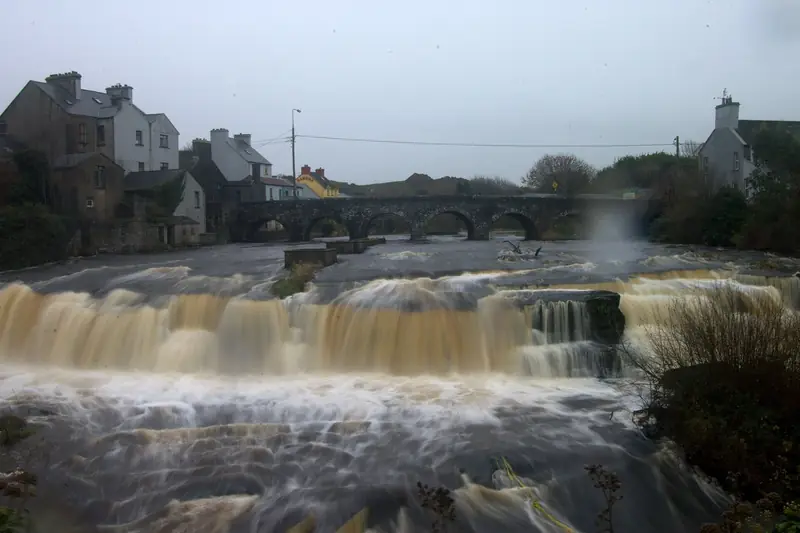 The Cascades on the river at Ennistymon in flood: brown, peaty water pouring over wide stone steps in front of an old arched bridge and the houses of the town.