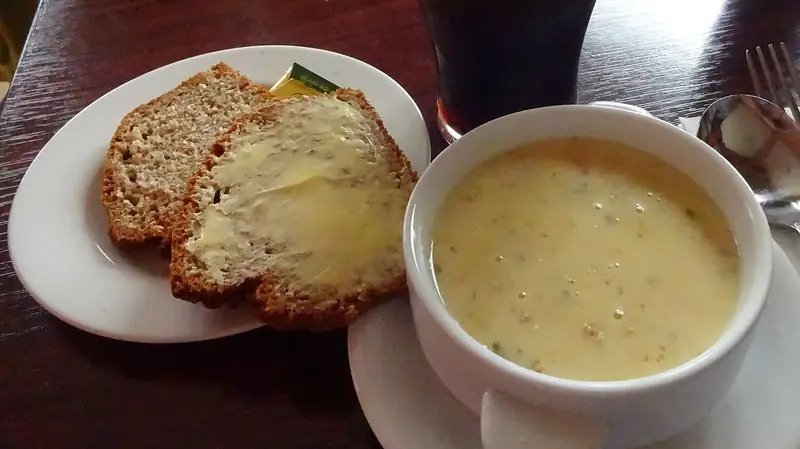 A white bowl of creamy seafood chowder beside two slices of buttered brown bread on a dark wooden table.