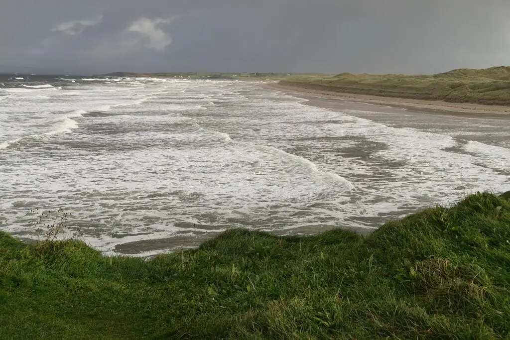 Grey Atlantic swell rolling in across Tullan Strand, with sand dunes behind and wind-flattened grass in the foreground.